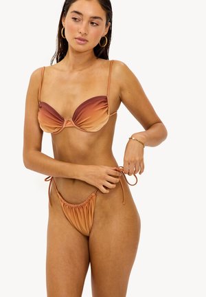 Femme ajustant les liens d'un bikini dégradé beige et marron, portant des boucles d'oreilles anneau dorées et un bracelet, sur fond blanc.