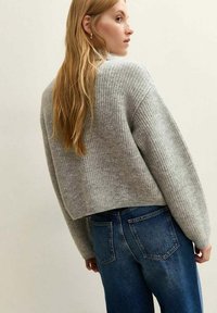 Woman with long blonde hair wearing a loose gray ribbed sweater and blue denim jeans, facing sideways against a plain background.