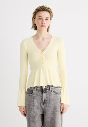 Kvinde iført en lysegul, knappet cardigan med udvidede manchetter og peplumkant, kombineret med gråvaske jeans med højtaljet pasform.