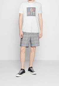 Camiseta blanca con un estampado geométrico multicolor sobre un fondo cuadrado, combinada con pantalones cortos de cuadros grises y zapatillas negras.