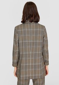 Hnědo-krémový kostkovaný blazer s jediným rozparkem na zadní straně, přiléhavý střih a dlouhé rukávy, s klasickým klopovým límcem a strukturovanými rameny.