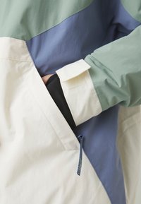 Coupe-vent en coloris mint, ivoire et bleu. Dispose d'une poche avant, de poignets de manche ajustables et d'un tissu texturé.