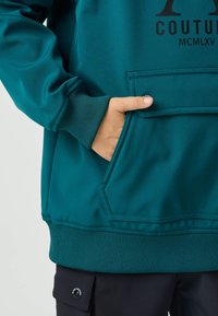 Felpa color teal con una grande tasca frontale, polsini e orlo a coste. Presenta una stampa del logo in nero e un piccolo dettaglio con bottone marchiato.