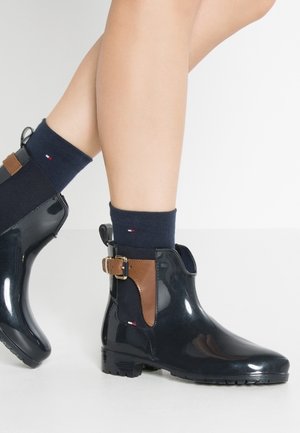 Jambes portant des bottines de pluie vernies noires à chevilles avec des accents de boucle marron et des chaussettes bleu marine ornées de petits logos rouges, blancs et bleus.