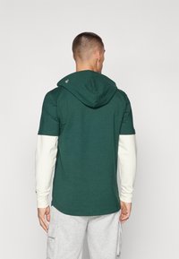 Zelená mikina s krátkym rukávom a bielou dlhým rukávom pod ňou, s nenápadným vyšívaným logom na kapucni a voľným strihom.