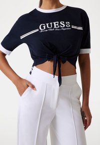 Navy cropped t-shirt with white trim and knot detail at the front, featuring "GUESS Sports Club Los Angeles" logo in white lettering. White high-waisted pants.