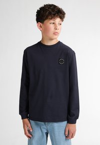 T-shirt a maniche lunghe blu marina realizzato in cotone, con collo rotondo e un piccolo patch decorativo sul petto, abbinato a jeans azzurri chiari.