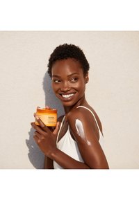 Orange burk med AHAVA kroppskräm hålls av en person, med vit kräm applicerad på deras arm. Bakgrunden är en ljus texturerad vägg.