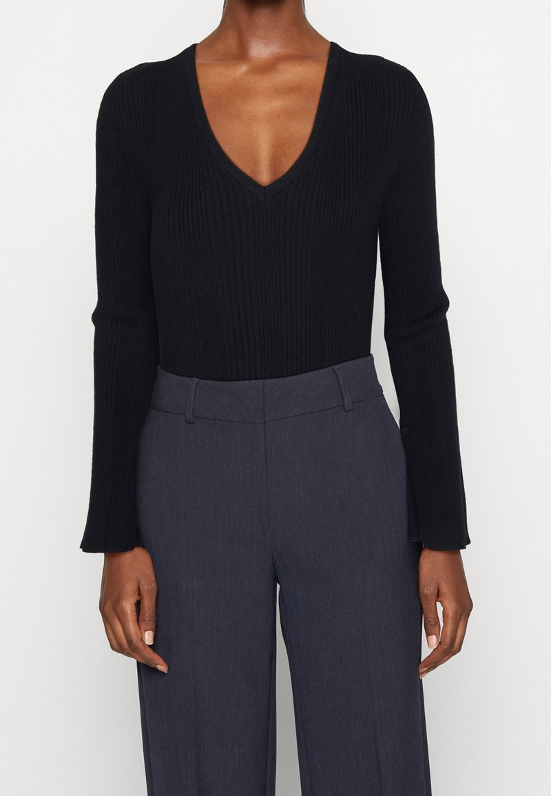 Femme portant un pull noir côtelé à manches longues et col en V, rentré dans un pantalon taille haute sur mesure gris foncé, debout devant un fond blanc.