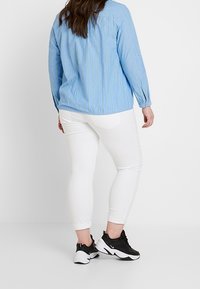 Hellblaue, gestreifte Bluse mit langen Ärmeln und lockerem Schnitt, kombiniert mit weißen Skinny Jeans und schwarzen Sportschuhen.