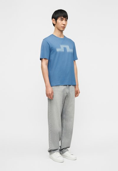 Μπλε κοντομάνικη μπλούζα με γραφικό σε σκιασμένο σχέδιο, συνδυασμένη με ανοιχτό γκρι τζιν σε άνετη εφαρμογή και λευκά αθλητικά παπούτσια.
