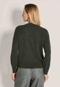 Frau mit blonden Haaren, die einen langärmeligen, dunkelgrauen Pullover und graue Nadelstreifen-Hosen trägt, von hinten vor einem schlichten Hintergrund betrachtet.