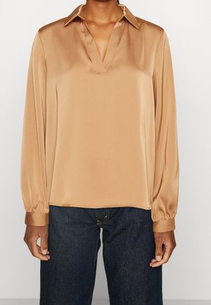 Henkilö, jolla on päällään pitkähihaisen, beige satiinipaidan, jossa on kaulus ja V-aukko, yhdistettynä tummansinisiin farkkuihin.