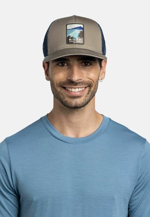 Lächelnder Mann mit kurzem dunklem Bart, der eine beige und marineblaue Kappe mit See- und Berglogo und ein hellblaues T-Shirt mit Rundhalsausschnitt trägt.