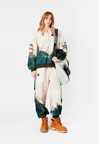 Conjunto de dos piezas que incluye una sudadera con capucha beige y azul verdoso con cremallera y estampado de montañas, combinada con pantalones deportivos a juego y botas naranjas.