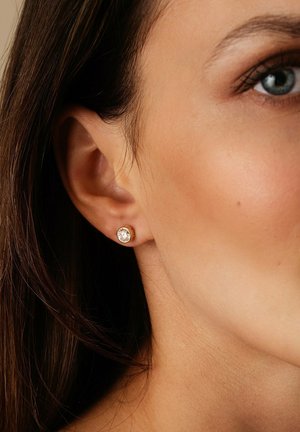 Nahaufnahme eines Ohrs einer Frau mit dunklem Haar, die einen kleinen, runden goldenen Ohrstecker mit einem klaren Edelstein trägt.