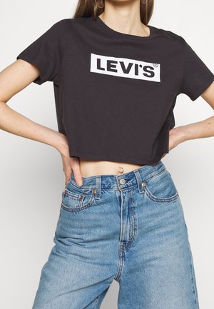 Femme portant un t-shirt noir court Levi's et un jean bleu taille haute avec un piercing visible au nombril, mains posées sur les hanches.