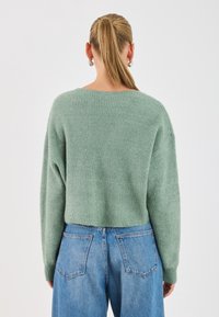 Pull vert tricoté à coupe décontractée, col bateau et manches longues, associé à un jean en denim bleu taille haute. La texture est douce et côtelée.