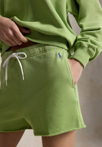 Osoba ubrana w jasnooliwkową bluzę z kapturem i dopasowane szorty z białym sznurkiem oraz małym niebieskim haftowanym logo po lewej stronie.