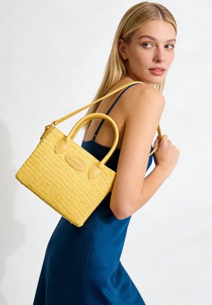Donna bionda in vestito blu che porta una borsa gialla intrecciata con doppi manici e tracolla, guardando indietro oltre la spalla.