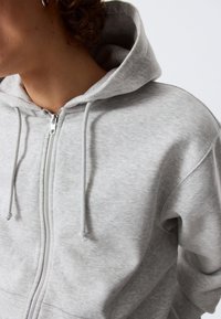 Sweat à capuche gris zippé en mélange de coton doux. Doté d'une grande capuche, de cordons de serrage et d'une texture lisse. La fermeture éclair descend sur le devant.