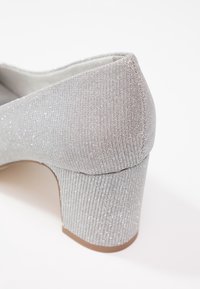 Gros plan d’une chaussure à talon bloc recouverte de tissu argenté avec un discret effet pailleté et un talon arrière arrondi.