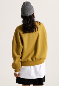 Overmaatse mosterdkleurige sweater met geribde boorden en zoom, gelaagd over een wit shirt en een zwarte rok. Grijze gebreide muts op het hoofd.