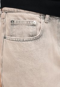 Κοντινό πλάνο στην τσέπη τζιν ανοιχτού μπεζ χρώματος με ασημένιες τάπες, φορεμένη με μαύρο τοπ με ριμπ ζώνη μέσης.