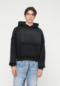 Črn oversize pulover s kapuco, kengursko žepom, raztrganim robom in vtisnjenim napisom "ATELIER". V kombinaciji z uničenimi svetlo modrimi denim kratkimi hlačami.