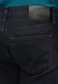 Tmavé džínsy s hladkou textúrou, ozdobené koženou nášivkou Calvin Klein na páse a jedným zadným vreckom s upraveným prešívaním.