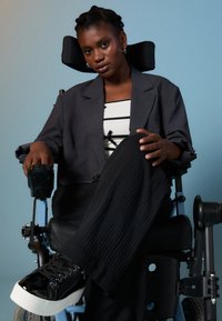 Άτομο με γκρι υπερμεγέθη σακάκι πάνω από μπλούζα με μαύρες και άσπρες ρίγες, καθισμένο σε μπλε αναπηρική καρέκλα, που φορά μαύρα sneakers με ύψος.