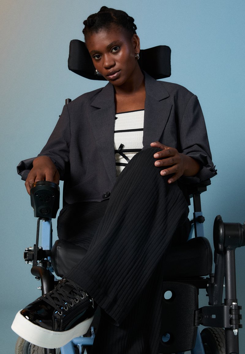 Άτομο με γκρι υπερμεγέθη σακάκι πάνω από μπλούζα με μαύρ�ες και άσπρες ρίγες, καθισμένο σε μπλε αναπηρική καρέκλα, που φορά μαύρα sneakers με ύψος.