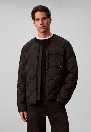 Mladý muž nosí černou prošívanou bundu s náprsní kapsou a logem Calvin Klein, v kombinaci s černými kalhotami a černo-bílou vrstvenou košilí.