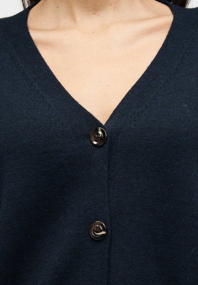 Ζακέτα σε ναυτικό μπλε με V-λαιμόκοψη, κατασκευασμένη από πλεκτό υλικό, με ένα μόνο μαύρο κουμπί στο μπροστινό μέρος και υφή υφάσματος.