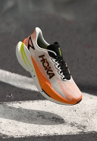 Sapatilha de corrida HOKA em malha branca com detalhes em laranja e amarelo, apresentando um logotipo preto, design leve e superior texturizado para respirabilidade.