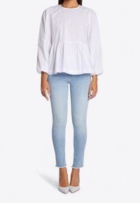 Donna con una blusa lunga bianca ampia a maniche lunghe, jeans attillati azzurri con orli sfilacciati e décolleté bianchi a punta su uno sfondo bianco.