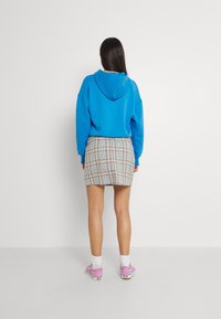 Sininen puuvillahuppari, jossa on huppu, yhdistettynä kuvioituun vaaleanbeigeen hameeseen, jossa on monivärisiä ruutuja. Mallilla on päällään vaaleanpunaiset lenkkarit valkoisten sukkien kanssa.