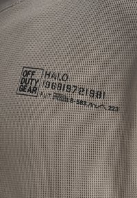 Waffelgestrickter beiger Stoff mit schwarzem Text „OFF DUTY GEAR“, „HALO“ und einer Folge von Zahlen und Buchstaben in einer lässigen Schriftart.