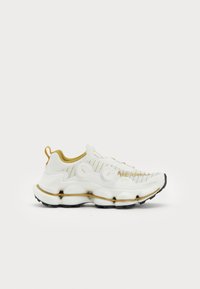Fehér sneaker textúrált felsőrésszel, arany díszítésekkel, két kör alakú fémzárral, vastag talppal, amelynek mintázata van, és saroknál egy húzószalaggal.