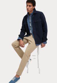 Giacca navy sopra una camicia a righe azzurre, abbinata a pantaloni beige e sneakers blu. Il modello è seduto su uno sgabello bianco.