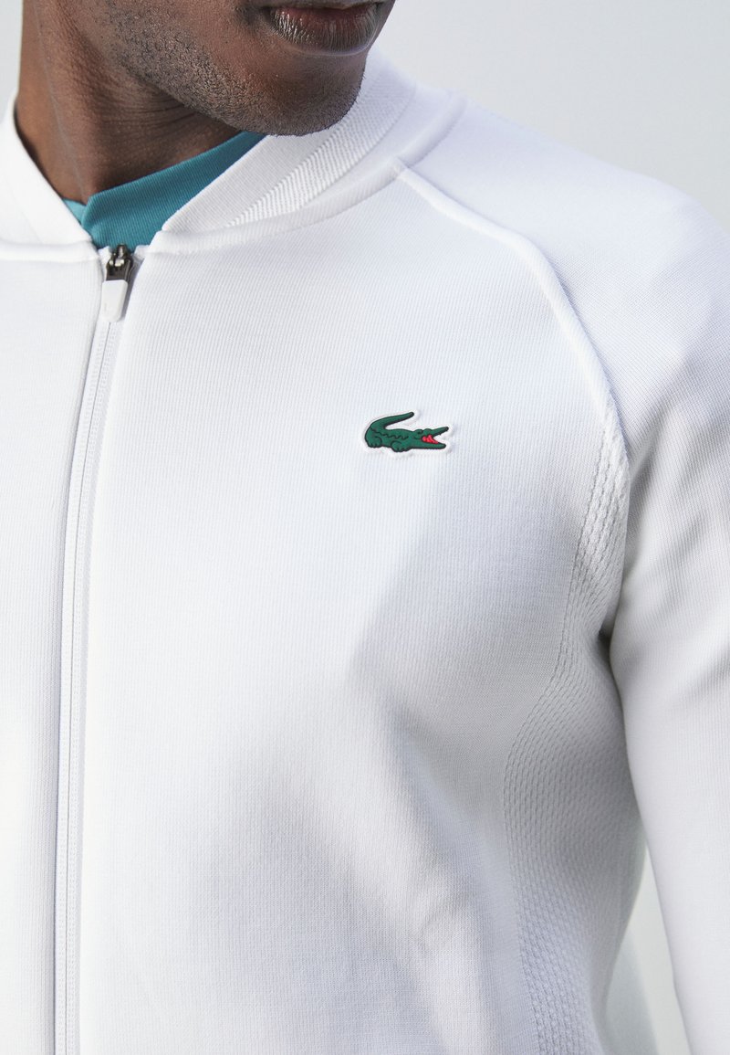 Veste zippée blanche en tissu texturé, arborant un logo de crocodile vert sur la poitrine. Col côtelé et coutures d'épaules.