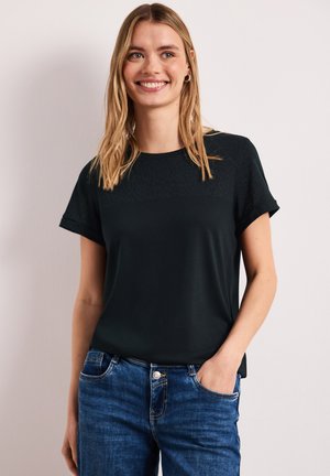 Ung kvinde smiler, iført en ensfarvet sort kortærmet skjorte og blå jeans, stående med den ene hånd i lommen mod en lys baggrund.