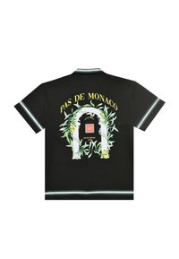 Svart kortermet skjorte med teksten "PAS DE MONACO" over en hvit bue innrammet med grønne blader og gule sitroner, med dekorativ kant på ermer og nederkant.