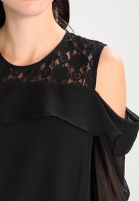 Blusa nera con un corpetto in pizzo, scollatura a volant e maniche a freddo trasparenti. Tessuto liscio, design elegante e motivo intricato.