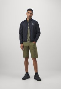 Schwarze leichte Jacke mit einem Frontreißverschluss, olivgrünes T-Shirt darunter und khakifarbene Shorts. Schwarze Sportschuhe runden das Outfit ab.