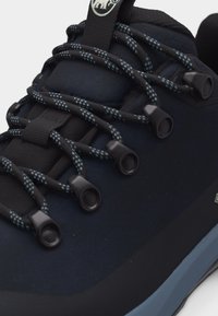 Prim-plan cu o sneaker albastru închis, cu șireturi rotunde negre și albastre cu model, trecute prin găuri negre și o limbă căptușită.
