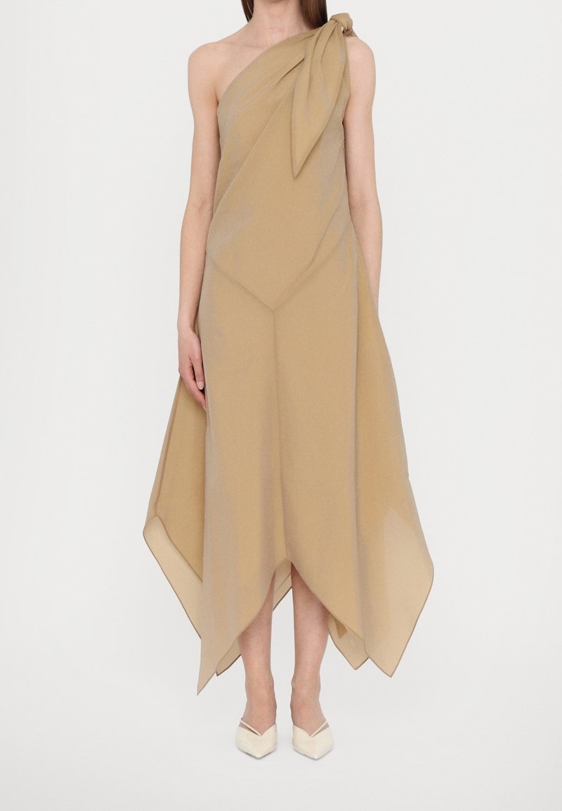 Femme portant une robe beige à une épaule avec un ourlet asymétrique et un détail noué, associée à des talons blancs à bouts pointus.