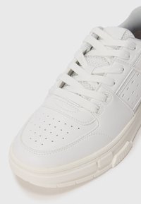 White sneaker featuring perforated toe, textured details on sides, flat white laces, and a thick rubber sole. Smooth synthetic material.