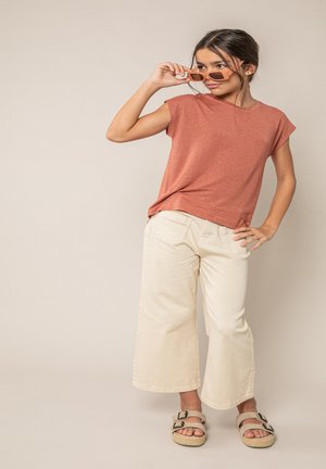 Jeune femme posant, portant un haut à manches courtes rouille, un pantalon large crème, des sandales beiges, et ajustant des lunettes de soleil rectangulaires orange.