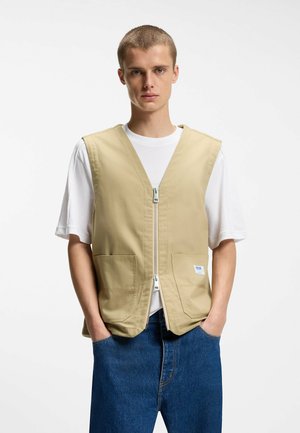 Giovane uomo con indosso un gilet beige con zip sopra una T-shirt bianca e jeans blu, in piedi con le mani in tasca davanti a uno sfondo bianco.